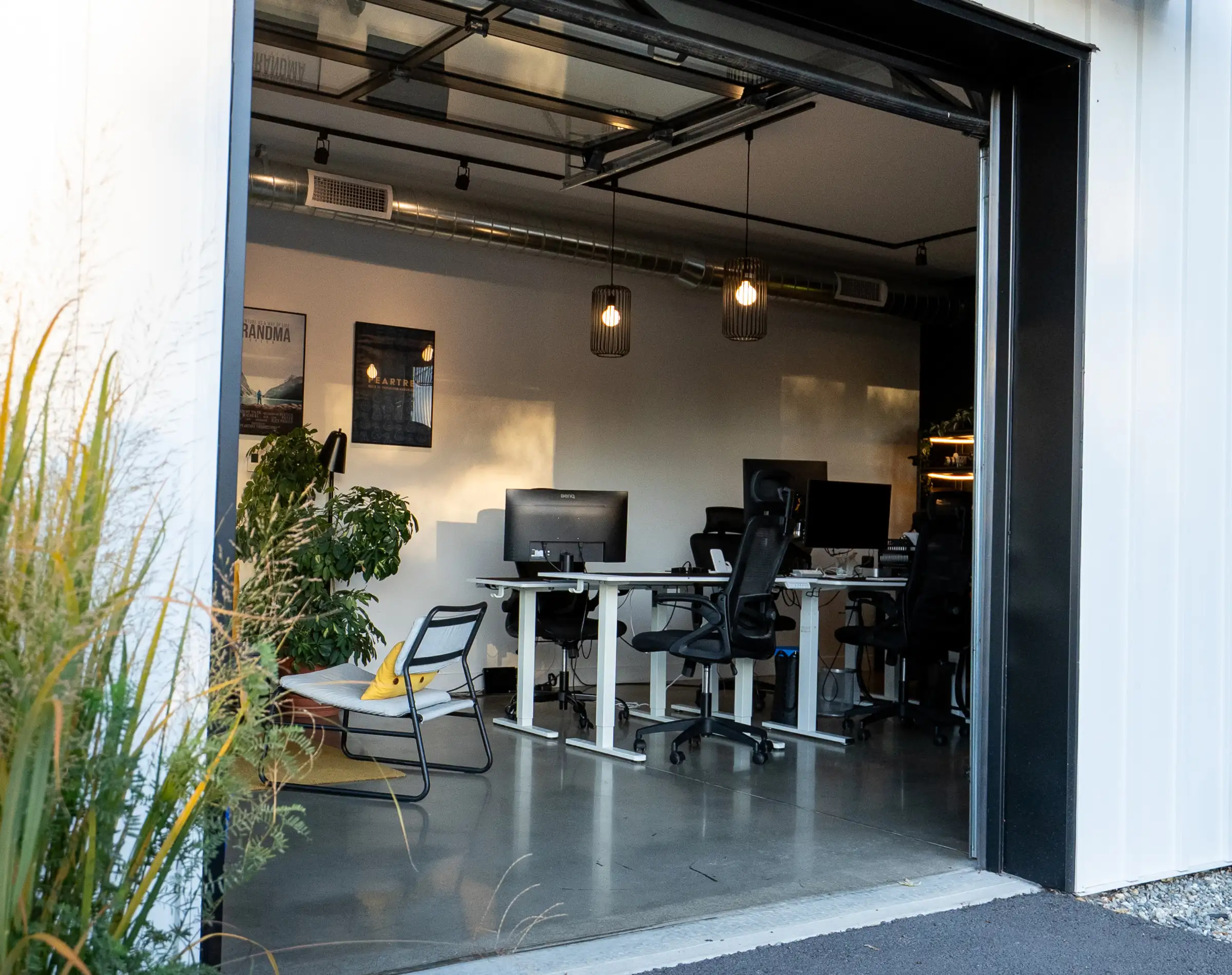 Bureaux de coworking vus par la porte de garage ouverte, avec postes de travail, plantes et suspensions lumineuses